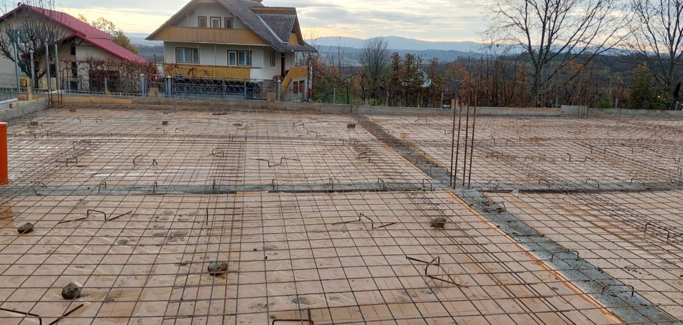 Fundație cu plasă de armătură la asfințit, cu casă tradițională galbenă și munți în fundal, Maramureș