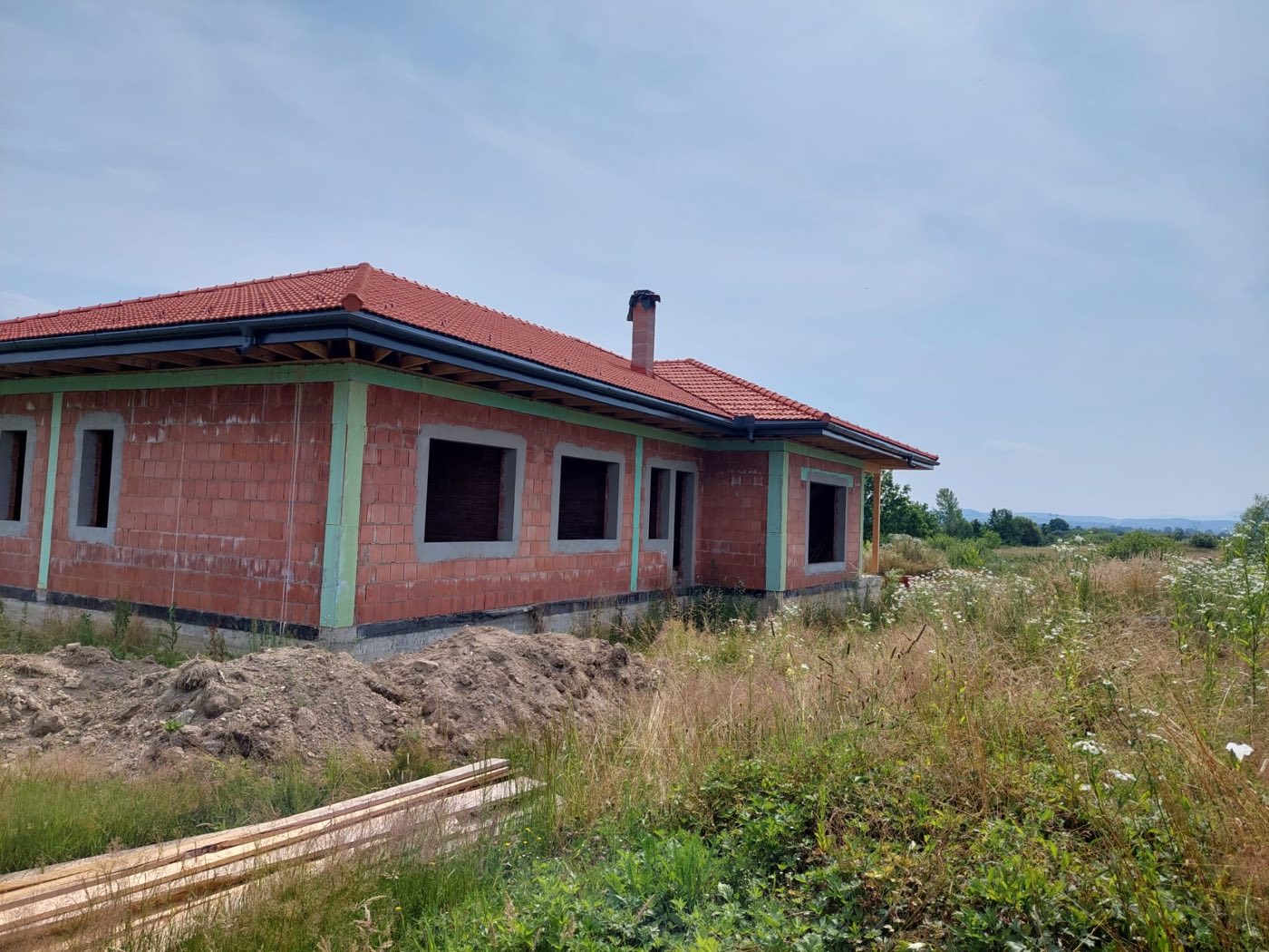 Casă finalizată cu acoperiș roșu și tâmplărie verde, într-o poiană cu flori