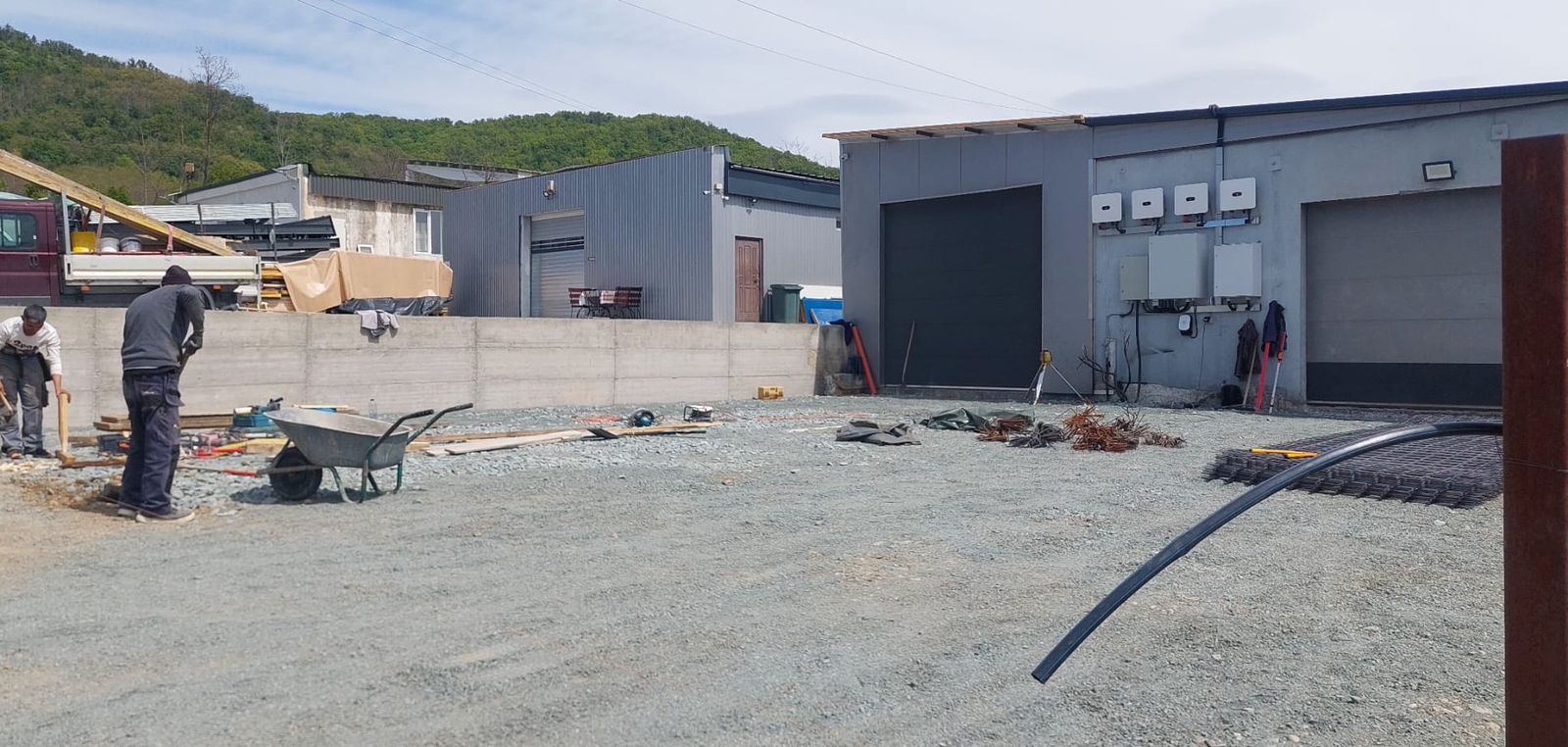 Șantier cu roabă și fier-beton pregătit, lângă o hală industrială