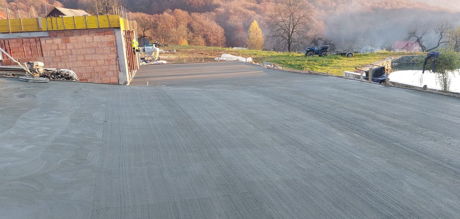 Muncitor nivelând beton lângă un zid de cărămidă, cu munți în ceață de toamnă