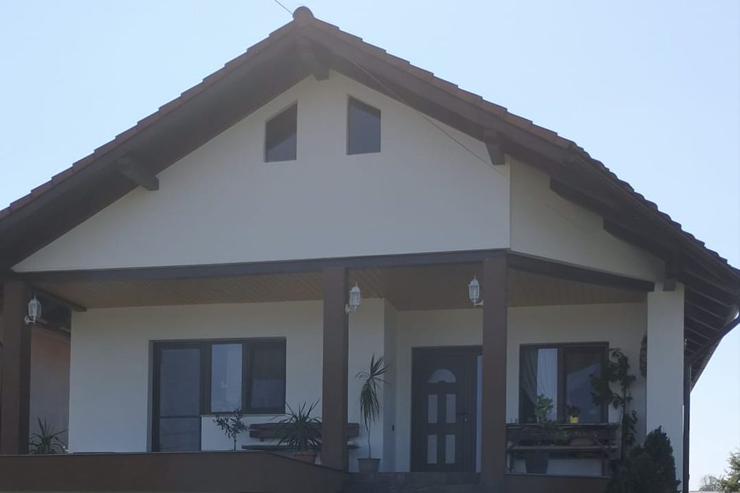 Aceeași casă finalizată: fațadă tencuită în alb, învelitoare din țiglă, tâmplărie montată, terasă acoperită cu felinare și scări din piatră.