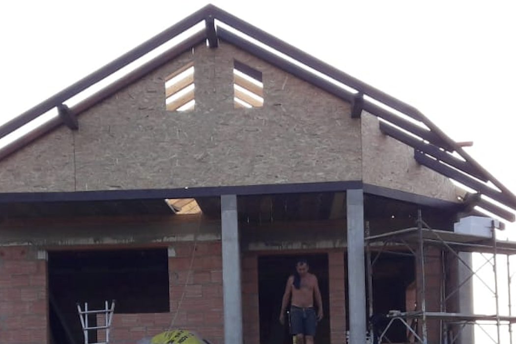 Casa la roșu: zidărie de cărămidă, fronton placat cu OSB, stâlpi de beton și schelă la fațadă.